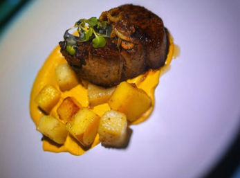 Gourmet seared beef medallion topped with microgreens, plated on a smear of bright orange purée with golden roasted potato cubes on a white plate — fine dining entrée.