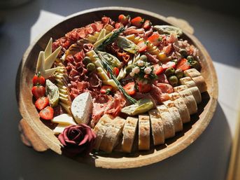Sunlit round wooden charcuterie board with sliced baguette, assorted cheeses (creamy wheel and wedges), folded cured meats, green olives, artichoke hearts, halved strawberries, rosemary sprig and edible flower garnish.
