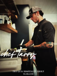 Tattooed chef in black uniform and cap using a spatula to cook in a busy restaurant kitchen