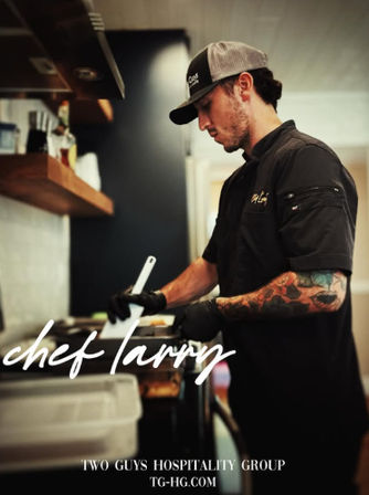 Tattooed chef in black uniform and cap using a spatula to cook in a busy restaurant kitchen