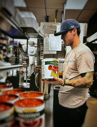 Tattooed cook in a busy commercial kitchen holding a large can of crushed tomatoes over an industrial mixer, with open cans of tomato sauce on a stainless counter.