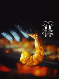 Close-up of a gloved hand holding a glazed grilled shrimp above glowing barbecue coals, golden sauce glistening with smoky orange bokeh in the background.