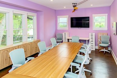 Bright meeting room with lavender walls, long wooden conference table, pastel blue ergonomic chairs, wall-mounted flat-screen TV, storage cabinets and large windows with natural light