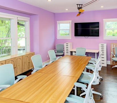 Bright meeting room with lavender walls, long wooden conference table, pastel blue ergonomic chairs, wall-mounted flat-screen TV, storage cabinets and large windows with natural light