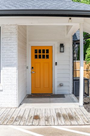 Bright yellow front door with small window panes in a white-sided home entryway, wood plank porch, outdoor lantern and welcome mat