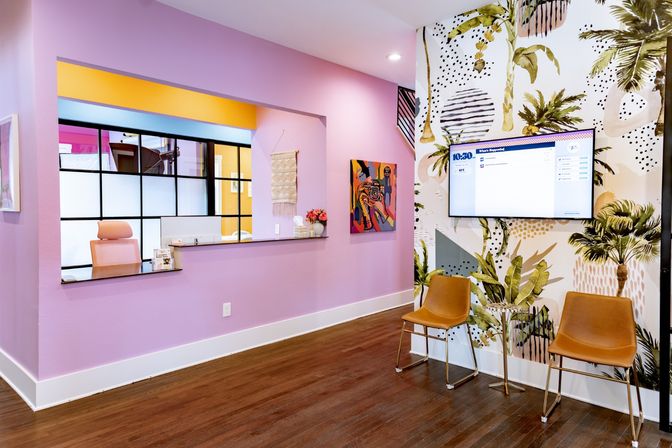 Colorful modern reception area with pastel pink walls, a service window, tropical palm mural, wall-mounted TV, and two tan waiting chairs on dark wood floors.
