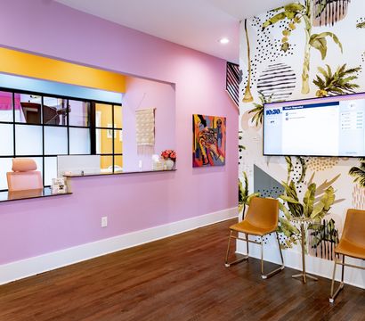 Colorful modern reception area with pastel pink walls, a service window, tropical palm mural, wall-mounted TV, and two tan waiting chairs on dark wood floors.
