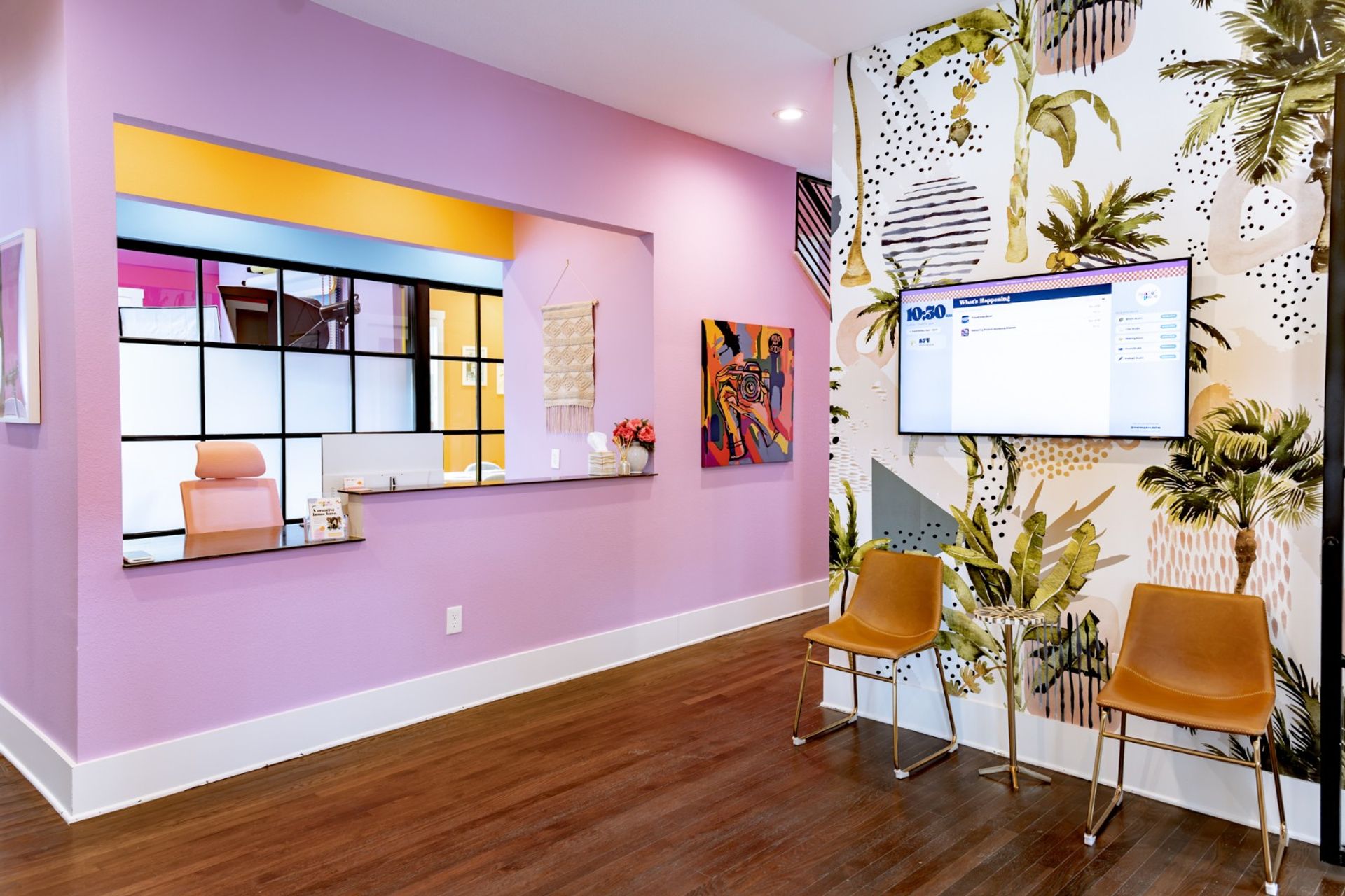 Colorful modern reception area with pastel pink walls, a service window, tropical palm mural, wall-mounted TV, and two tan waiting chairs on dark wood floors.