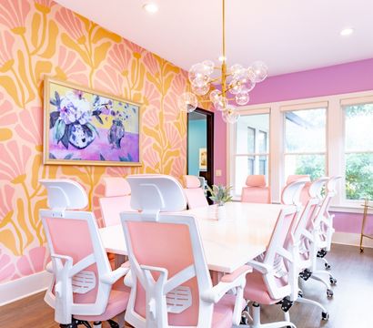 Sunlit boutique conference room with pink ergonomic chairs, white table, bold pink-and-gold floral wallpaper, large windows and bubble chandelier.