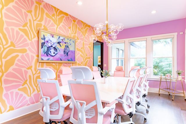 Sunlit boutique conference room with pink ergonomic chairs, white table, bold pink-and-gold floral wallpaper, large windows and bubble chandelier.