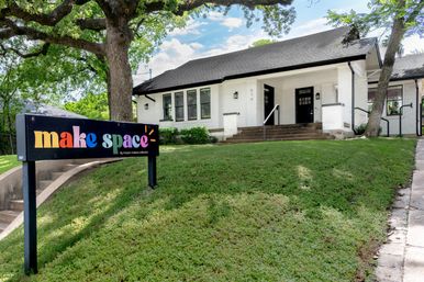 Charming white brick bungalow with wide front steps, giant oak tree and a colorful yard sign marking a community creative space on a sloped green lawn