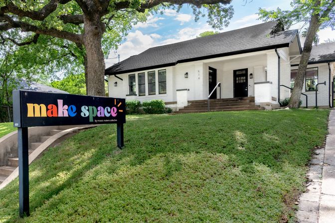 Charming white brick bungalow with wide front steps, giant oak tree and a colorful yard sign marking a community creative space on a sloped green lawn