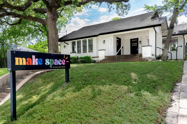 Charming white brick bungalow with wide front steps, giant oak tree and a colorful yard sign marking a community creative space on a sloped green lawn