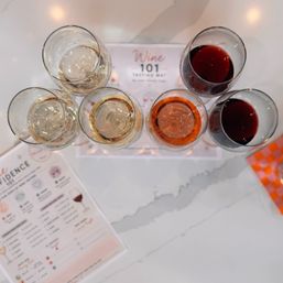 Overhead shot of a six-glass wine flight on a 'Wine 101' tasting mat—pale whites, a rosé and two reds—on a white marble tabletop with a tasting notes sheet at the side.