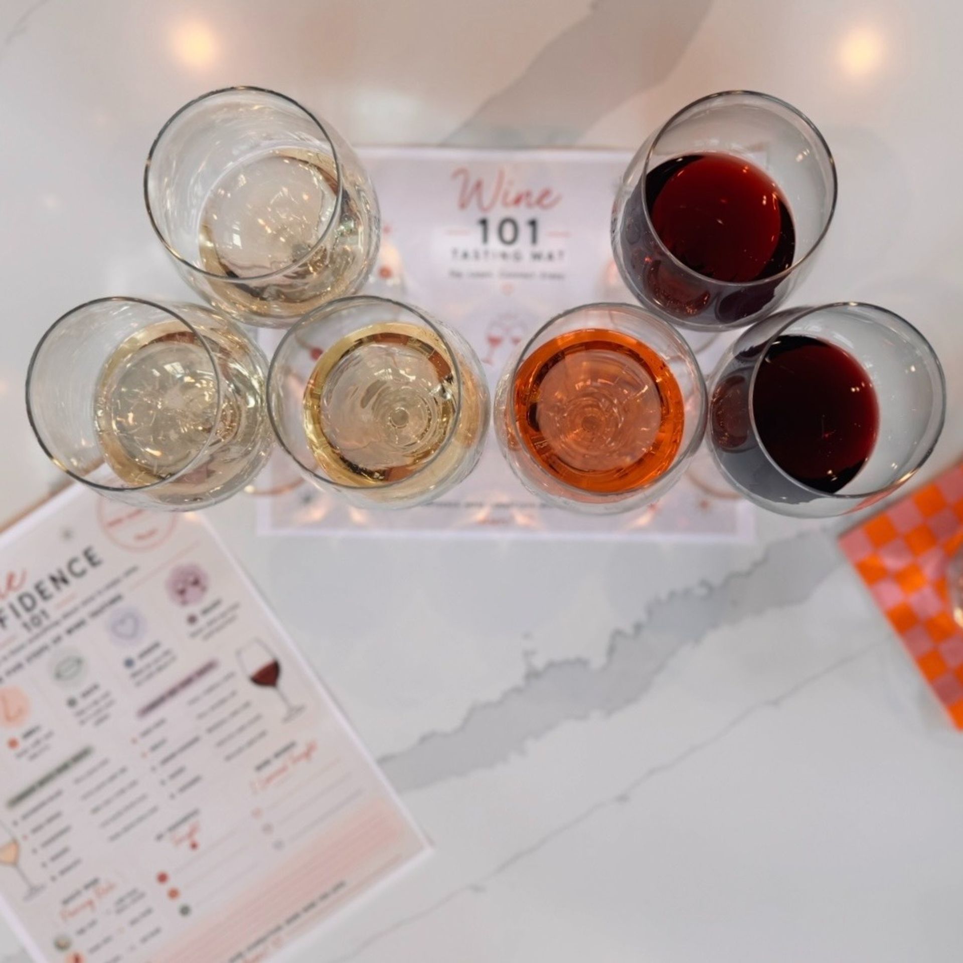 Overhead shot of a six-glass wine flight on a 'Wine 101' tasting mat—pale whites, a rosé and two reds—on a white marble tabletop with a tasting notes sheet at the side.