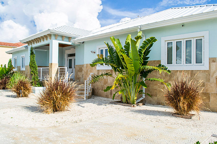 Sunny pastel-blue coastal cottage with white metal roof, stone lower facade, sandy yard and tropical landscaping of banana palms and ornamental grasses.