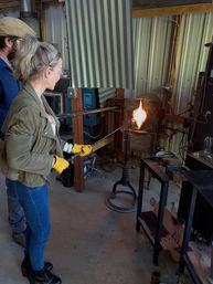 Hands-on glassblowing: person in safety glasses and yellow gloves shaping glowing molten glass at a studio furnace.