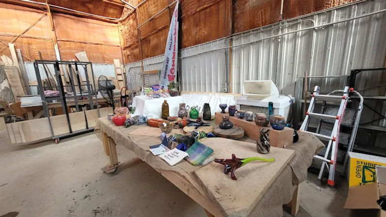Rustic warehouse artist studio with burlap-covered tables displaying colorful hand-blown glass bowls, vases and sculptures, surrounded by ladders, racks and workshop tools — artisan glass display in a workshop setting.