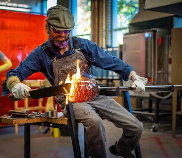 Glassblower shaping glowing molten glass on a punty rod in an indoor studio workshop, sparks flying as the furnace-heated art piece is formed.