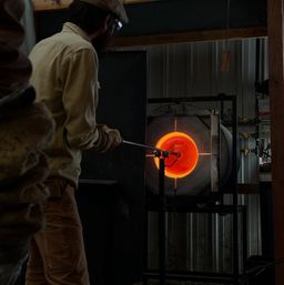 Artisan glassblower in a workshop heating a glowing orange orb of molten glass in a furnace and shaping it with a blowpipe.