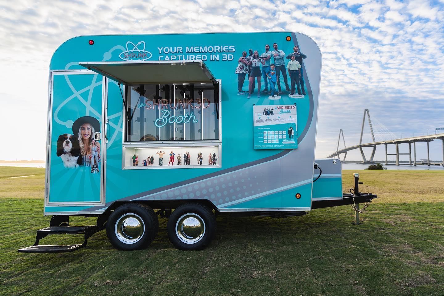 Teal mobile 3D-photo trailer parked on a grassy waterfront park with an open service window showing miniature family figurines, a cable-stayed bridge and blue cloudy sky in the background.