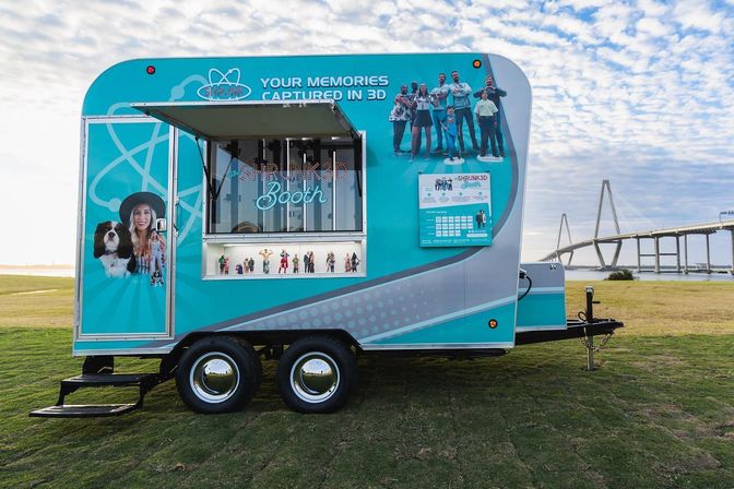 Teal mobile 3D-photo trailer parked on a grassy waterfront park with an open service window showing miniature family figurines, a cable-stayed bridge and blue cloudy sky in the background.