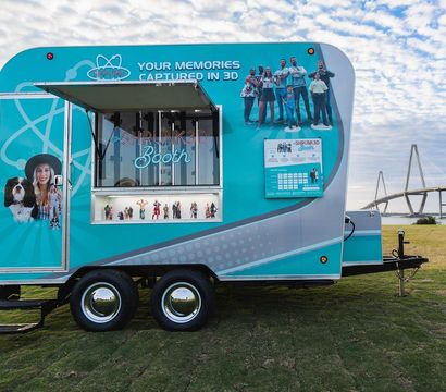 Teal mobile 3D-photo trailer parked on a grassy waterfront park with an open service window showing miniature family figurines, a cable-stayed bridge and blue cloudy sky in the background.