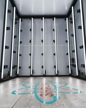 Sleek empty LED-lit photo studio booth with vertical white light bars on gray walls and a wood-look floor marked by a turquoise graphic logo — modern indoor product and portrait lightbox.