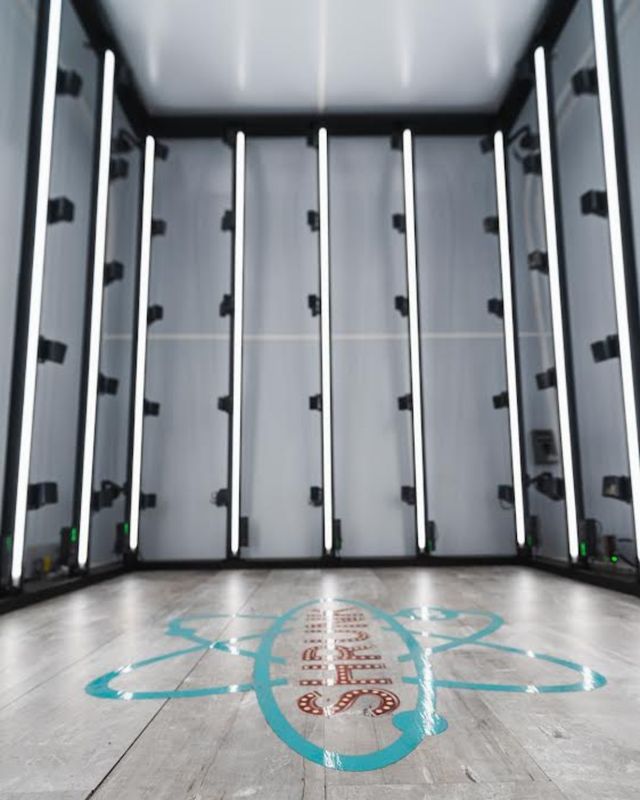 Sleek empty LED-lit photo studio booth with vertical white light bars on gray walls and a wood-look floor marked by a turquoise graphic logo — modern indoor product and portrait lightbox.