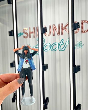 Hand holding a tiny 3D-printed miniature of a playful person in a denim vest and hoodie, tongue-out pose against a vertical LED-lit photo booth backdrop