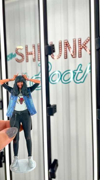 Hand holding a tiny 3D-printed miniature of a playful person in a denim vest and hoodie, tongue-out pose against a vertical LED-lit photo booth backdrop