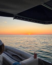 Sunset over calm ocean seen from a boat under a dark canopy — warm orange-pink sky with the sun dipping at the horizon, rippling water reflecting color, cushioned seating and a cup in a cupholder visible.