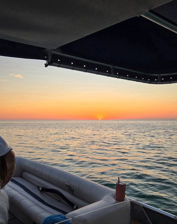 Sunset over calm ocean seen from a boat under a dark canopy — warm orange-pink sky with the sun dipping at the horizon, rippling water reflecting color, cushioned seating and a cup in a cupholder visible.
