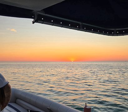 Sunset over calm ocean seen from a boat under a dark canopy — warm orange-pink sky with the sun dipping at the horizon, rippling water reflecting color, cushioned seating and a cup in a cupholder visible.