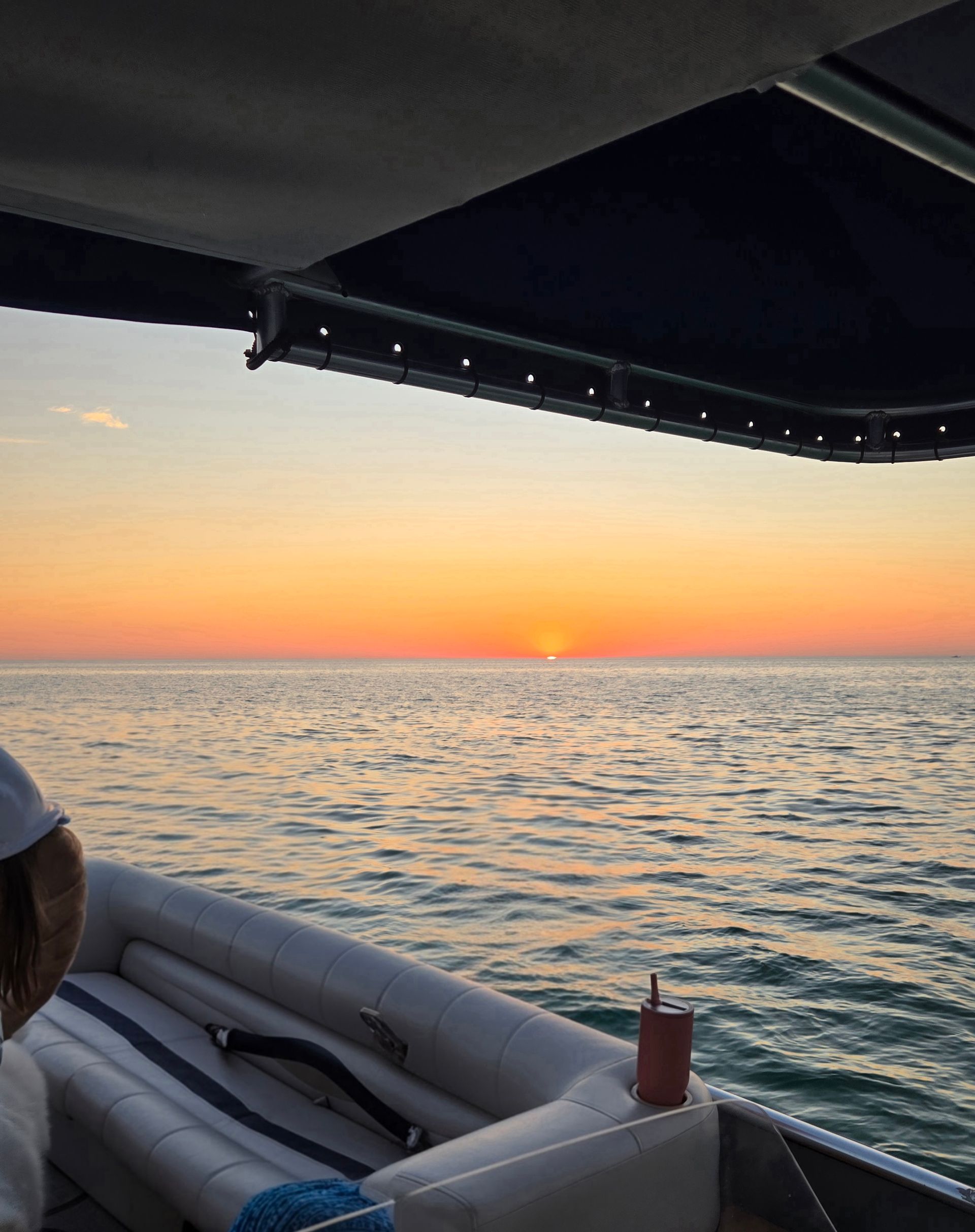 Sunset over calm ocean seen from a boat under a dark canopy — warm orange-pink sky with the sun dipping at the horizon, rippling water reflecting color, cushioned seating and a cup in a cupholder visible.