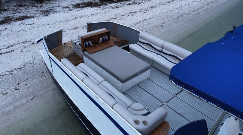 Pontoon boat moored at a sandy shore with white-and-navy cushioned seating, a central gray sunbed, wooden decking and a blue canopy over clear shallow water.