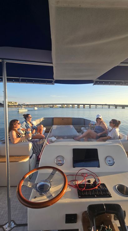 Friends sipping drinks and relaxing on a pontoon boat at sunset, wooden steering wheel and controls in the foreground, calm bay waters with small boats and a long bridge on the horizon