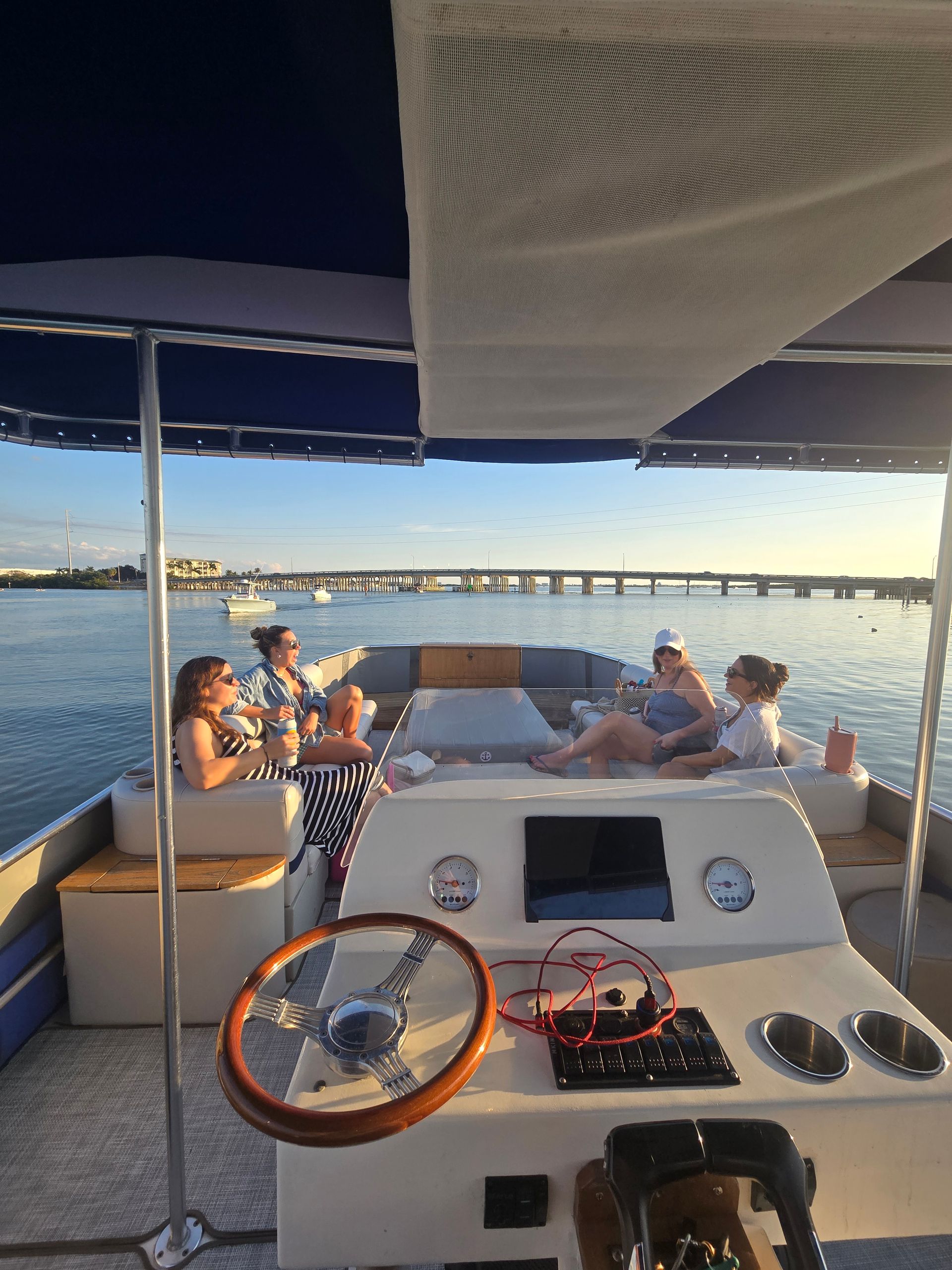Friends sipping drinks and relaxing on a pontoon boat at sunset, wooden steering wheel and controls in the foreground, calm bay waters with small boats and a long bridge on the horizon