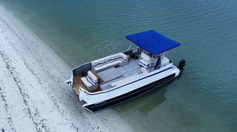 Aerial view of a white pontoon boat with a bright blue canopy anchored in shallow turquoise water along a white sandy beach, relaxed coastal scene