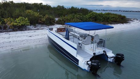 Blue-canopied pontoon boat anchored in a shallow turquoise bay beside a white sandy beach and mangrove-covered shoreline, coastal homes visible across the water.