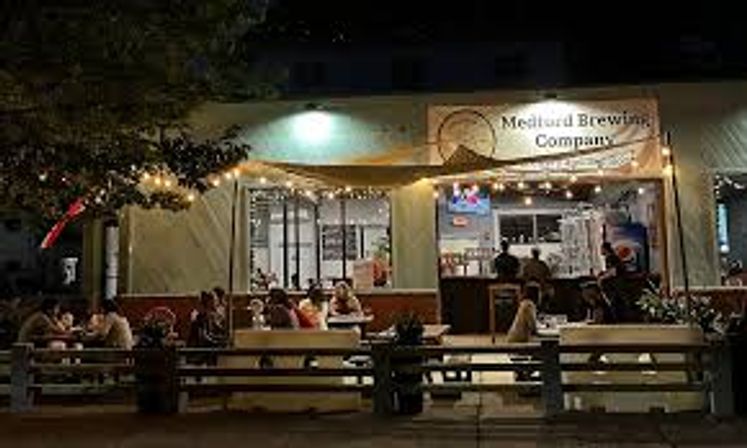 Nighttime street view of a lively brewpub patio with string lights, outdoor dining benches and tables filled with diners, open service window and glowing interior