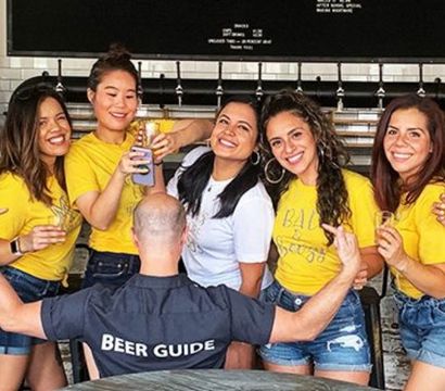 Group of friends in matching yellow tees and denim shorts enjoying beer tasting at a craft brewery taproom while a guide wearing a 'Beer Guide' shirt faces them with arms raised.