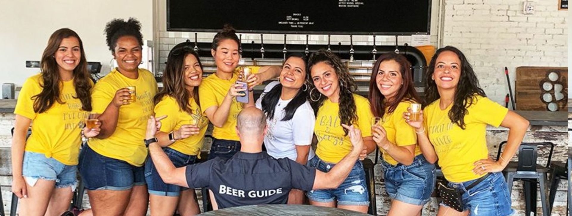 Group of friends in matching yellow tees and denim shorts enjoying beer tasting at a craft brewery taproom while a guide wearing a 'Beer Guide' shirt faces them with arms raised.