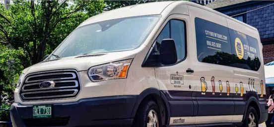 White passenger van with brewery-themed beer glass graphics and dark windows parked on a tree-lined urban street