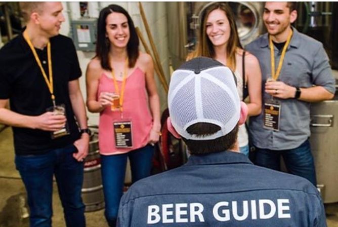 Beer guide in mesh cap speaks to a smiling group of four on a brewery tour, each holding tasting glasses and lanyards among stainless-steel tanks and kegs