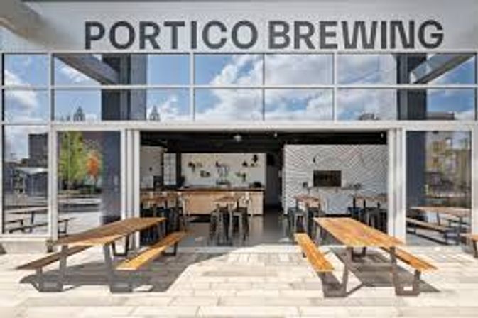 Sunny urban brewery patio with open glass garage doors, long wooden picnic tables, and an industrial taproom with bar stools and modern decor.
