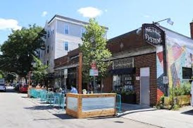 Sunny urban street with a wooden parklet and turquoise outdoor dining barriers, brick storefronts featuring a colorful mural and mixed-use apartments above.
