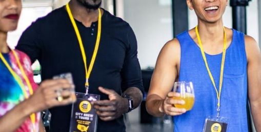 Group of adults on a brewery tour in a bright taproom, wearing yellow lanyards and event badges and holding glasses of craft beer, smiling and chatting.