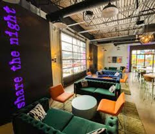 Bright industrial loft lounge with emerald velvet sofas, orange accent chairs, round coffee tables, neon purple wall sign, large garage-style windows and exposed ceiling ducts