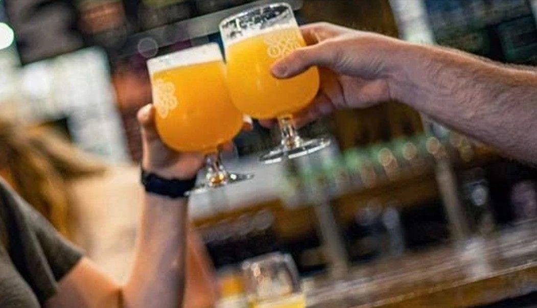 Two people clinking tulip glasses of hazy orange craft beer at a lively taproom — casual cheers